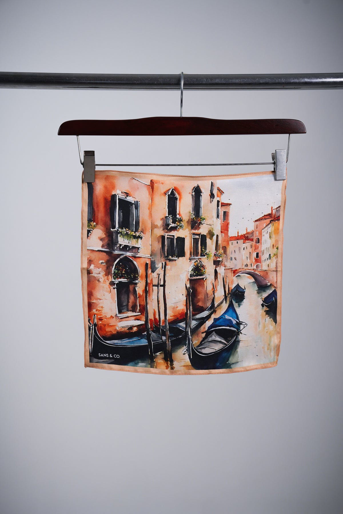 A square scarf featuring a watercolor-style painting of a Venice canal with gondolas and warm-toned buildings, hanging from a wooden hanger on a metal rod against a light gray background.
