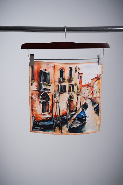 A square scarf featuring a watercolor-style painting of a Venice canal with gondolas and warm-toned buildings, hanging from a wooden hanger on a metal rod against a light gray background.
