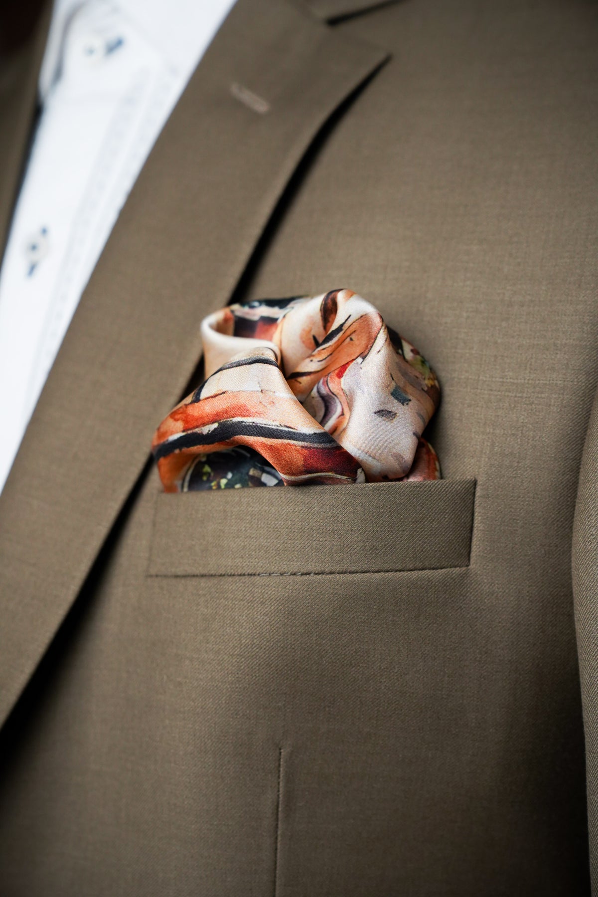 A close-up of a men's brown suit jacket with a patterned silk pocket square tucked into the breast pocket, layered over a white dress shirt.